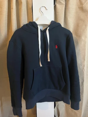 Ralph lauren hoodie med broderad logga - storlek M - Stilren mörkblå hoodie från Ralph Lauren i klassisk modell. Hoodien har en avslappnad passform med justerbar huva och en praktisk känguruficka framtill. Den ikoniska broderade loggan i rött på bröstet ger ett tidlöst och exklusivt uttryck.  ✔ Storlek: M men kort i storlek! ✔ Färg: Mörkblå ✔ Modell: Hoodie (utan dragkedja) ✔ Detaljer: Broderad logga på bröstet, känguruficka, ribbade muddar och nederkant, vita snören ✔ Material: Mjuk och bekväm kvalitet ✔ Skick: Väldigt bra skick – sparsamt använd