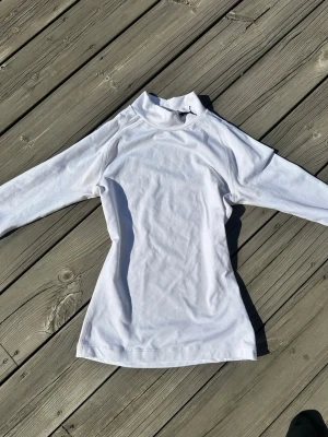 Vit långärmad träningströja Puma - Säljer en vit långärmad träningströja från Puma med hög krage och svart Puma-logga på halsen. Tillverkad i ett mjukt och stretchigt tyg som passar perfekt till träning eller sportaktiviteter.