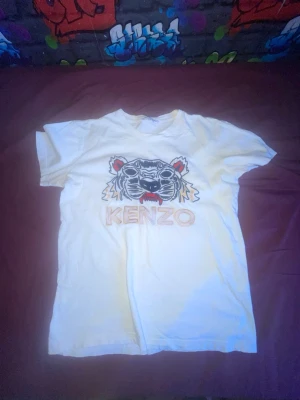 Vit Kenzo t-shirt med tigertryck - Säljer en vit t-shirt från Kenzo med det ikoniska tigerhuvudet broderat framtill och Kenzo-loggan under. T-shirten har korta ärmar och är gjord i mjuk bomull. Perfekt för dig som vill ha en statement tee med streetwear-vibe.