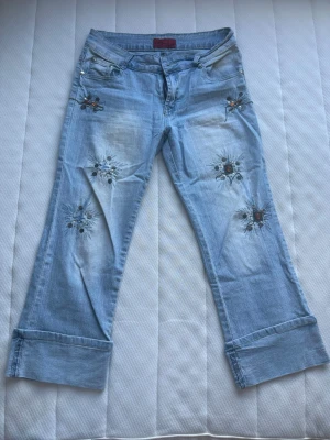 Ljusblå broderade jeans med vida ben - Unika ljusblå jeans med broderade blommor och pärldetaljer på låren och knäna. Jeansen har vida ben och uppvikta benslut för en avslappnad look. Perfekta för dig som vill sticka ut med coola detaljer och en trendig passform. Saknar två paletter
