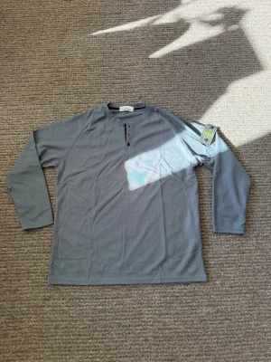 Grå långärmad tröja från Stone Island - Säljer en grå långärmad tröja från Stone Island med klassisk patch på vänster ärm. Tröjan har knappar vid halsen och är tillverkad i ett lätt, strukturerat material som ger en avslappnad men ändå stilren look.