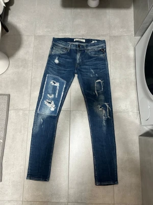 Blå slitna jeans från Replay - W31 L32. Säljer ett par blå  jeans från Replay med slitningar och patchdetaljer på benen. Jeansen har fem fickor, dragkedjegylf och klassisk Replay-lapp bak i midjan. Materialet är denim med stretch för skön passform.