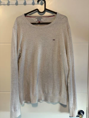 Ljusgrå stickad tröja Tommy Hilfiger - Ljusgrå stickad tröja från Tommy Hilfiger med rund halsringning och diskret logga på bröstet. Tröjan har långa ärmar och ribbade muddar vid ärmslut och nederkant. Perfekt basplagg för lager på lager och enkel att matcha.