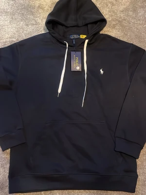 Marinblå hoodie från Polo Ralph Lauren - Snygg marinblå hoodie från Polo Ralph Lauren med vit broderad logga på bröstet och vita dragsnören i huvan. Klassisk känguruficka framtill och ribbade muddar vid ärmar och nederkant. Tillverkad i mjukt bomullsmaterial för skön känsla. Den har storlek L men passar som M. (Rep)