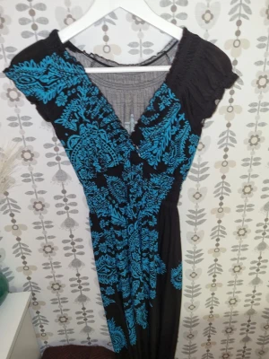 Svart långklänning med blått mönster - Svart långklänning med turkosblått blommigt mönster och omlott-ringning. Klänningen har korta ärmar och smockdetaljer upptill för en bekväm passform. Perfekt för dig som vill sticka ut med färg och mönster.