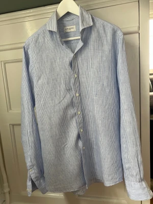 Randig linneskjorta från John Henric - Snygg blårandig linneskjorta från John Henric med klassisk krage och knappar framtill. Tillverkad i ett lätt och luftigt bomullstyg, perfekt för en stilren look. Långärmad modell med smala vita och blå ränder som ger en fräsch känsla. 