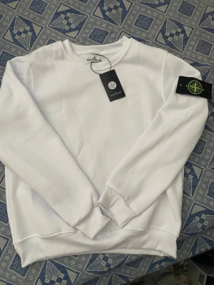 Vit sweatshirt från Stone Island - Säljer en clean vit sweatshirt från Stone Island med den klassiska svarta logotypen på ärmen. Tröjan har rund halsringning, ribbade muddar och är tillverkad i mjukt material. Perfekt för dig som gillar stilrena plagg med streetkänsla.