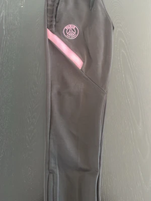 Svarta PSG träningsbyxor Nike - Snygga svarta träningsbyxor från Nike med PSG-logga och rosa detaljer. Byxorna har elastisk midja, dragkedja vid fickorna och en rosa Nike Swoosh på ena benet. Perfekta för träning eller chill. Materialet är mjukt och stretchigt för bästa komfort.