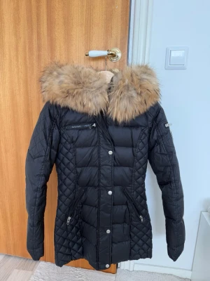 Svart dunjacka Rockandblue päls - Snygg svart dunjacka från Rockandblue med stor fluffig äkta päls på huvan. Jackan har quiltade detaljer, dragkedjor på fickorna och en patch på ärmen. Perfekt för kalla vinterdagar och riktigt trendig look.