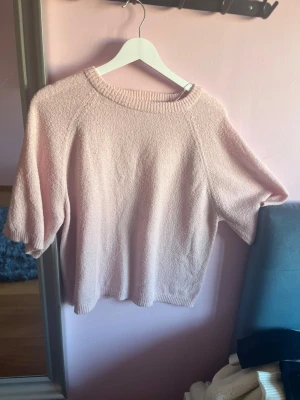 Ljusrosa stickad tröja med kort ärm - Söt ljusrosa stickad tröja med korta ärmar och rund halsringning. Tröjan har en enkel, avslappnad passform och ribbstickade detaljer vid hals och nederkant. Perfekt för lager på lager och en mjuk känsla.