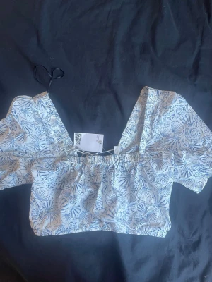 Blåvit blommig omlottopp med snörning - Supersöt omlottopp från H&M i blåvitt blommigt mönster. Toppen har korta puffärmar, snörning framtill och en croppad passform. Perfekt för dig som gillar en feminin och trendig stil. Materialet känns lätt och luftigt, troligen bomull.(klicka ej köp nu) kontakta gärna och Swish går ❤️