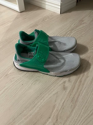 Nike Sock Dart grå/gröna sneakers - Nike Sock Dart sneakers i grått meshmaterial med grönt plastspänne och detaljer. Skon har en låg silhuett, vit sula med gröna inslag och en minimalistisk design utan klassiska snören. Perfekt för dig som gillar sportig och modern stil.