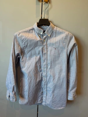 Blårandig skjorta från Gant - Klassisk blårandig skjorta från Gant med button down-krage och små broderade logodetaljer på bröstet. Skjortan är långärmad och tillverkad i mjuk bomull, perfekt för en clean och stilren look.