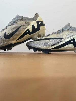 Nike Air Zoom Mercurial fotbollsskor - Snygga Nike Air Zoom Mercurial fotbollsskor i grått och beige med svart swoosh och AIR-text på sidan. Skorna har låg profil, snörning och en ovandel i lätt syntetmaterial för bra bollkontroll. Perfekta för dig som vill ha speed och stil på planen. Elite FG. Pris kan diskuteras.