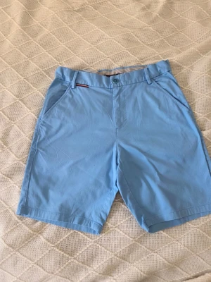 Ljusblå shorts från Tommy Hilfiger - Snygga ljusblå shorts från Tommy Hilfiger med klassisk design och raka ben. Shortsen har sidofickor, bälteshällor och knappstängning. Perfekta för varma dagar och har en stilren look med diskret logga vid fickan. 