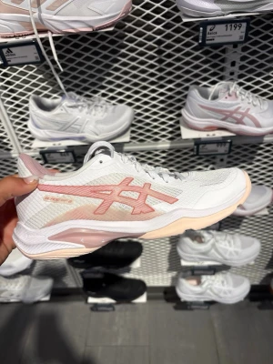Vita och rosa sneakers från Asics - Snygga vita sneakers från Asics med ljusrosa detaljer och logga på sidan. Skon har en låg profil, meshmaterial för bra andningsförmåga och en dämpad sula i rosa och vitt. Perfekt för dig som vill ha både stil och komfort till träningen eller vardagslooken.