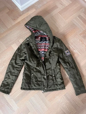 Olivgrön tunn jacka från bpc - Snygg olivgrön jacka från bpc med huva och mönstrat innerfoder i rött, svart och vitt. Jackan har dragkedja och knappar framtill samt två fickor. Perfekt för dig som vill ha en lätt och stilren jacka med coola detaljer.