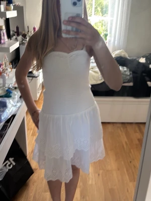 Vit axelbandslös sommarklänning - Söt vit axelbandslös klänning med smockad överdel och volangig kjol. Klänningen har broderade detaljer på kjolen och är perfekt för varma dagar. Tillverkad i lätt bomull som känns skön mot huden. Klänningen är helt ny och kostar 527 ungefär iaf något med 500 kr 