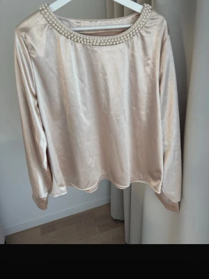 Champagnefärgad topp med pärlor från SHEIN storlek: XL - Snygg champagnefärgad långärmad topp med rund halsringning dekorerad med pärlor. Materialet har en lätt glansig satinlook som ger en lyxig känsla. Perfekt att styla med jeans eller kjol för en trendig look.