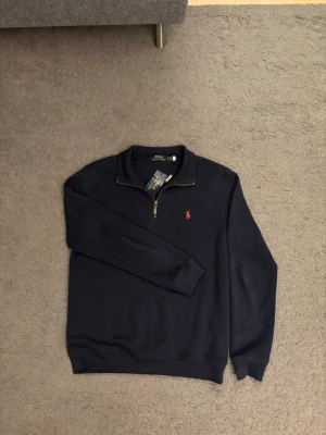Mörkblå half-zip tröja Polo Ralph Lauren - Mörkblå tröja från Polo Ralph Lauren med half-zip och klassisk röd broderad logga på bröstet. Tröjan har ribbade muddar och krage, samt mjukt bomullsmaterial som ger en clean och stilren look.