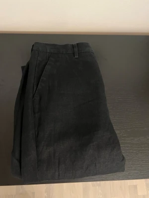 Svarta linne kostymbyxor - Säljer ett par svarta linnebyxor med raka ben och klassisk passform. Byxorna har sidofickor, bälteshällor och stängs med knapp och dragkedja. Perfekta för en avslappnad men stilren look.