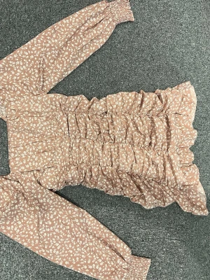 Blommig kortklänning med volang - Supersöt kortklänning i beige med vitt blommönster. Klänningen har långa ärmar med resår vid handlederna, volangdetaljer och smockad midja som ger en snygg passform. Perfekt för vår och sommar!