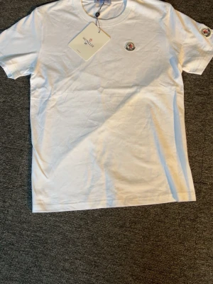 Vit Moncler t-shirt med logga - Säljer en stilren vit t-shirt från Moncler med klassisk logga på bröstet och ärmen. T-shirten har rund hals och korta ärmar. Tillverkad i mjuk bomull för en skön känsla. Perfekt till jeans eller shorts för en clean look.
