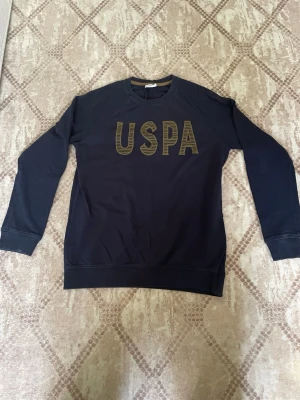 Svart sweatshirt från U.S. Polo Assn - Svart sweatshirt med stora gula USPA-bokstäver på bröstet. Tröjan har rund halsringning och långa ärmar med diskret textur på axlarna. Perfekt för en avslappnad och sportig stil.
