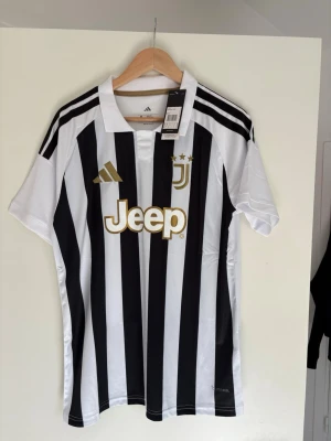 Juventus matchtröja Adidas svartvit M - Juventus officiella matchtröja från Adidas i svartvitt randigt med gulddetaljer och Jeep-logga på bröstet. Slim fit-modell med klassisk krage och tre stjärnor ovanför klubbmärket. Tillverkad i lätt och ventilerande material.
