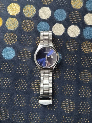 Silverfärgad klocka från Moretime - Stilren klocka från Moretime med silverfärgat metallarmband och mörkblå urtavla. Klockan har datumvisning och klassisk design som passar till det mesta. Länken är i metall och spännet är graverat med varumärket.