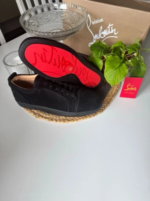Svarta Louboutin sneakers med röd sula - Snygga svarta sneakers från Christian Louboutin i mocka med klassisk röd sula och svarta skosnören. Låg modell med stilren design och exklusiv känsla. Perfekta för dig som vill sticka ut med en ikonisk detalj under skon.