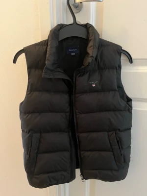 Svart dunväst från GANT - Snygg svart dunväst från GANT med hög krage och dragkedja framtill. Västen har två stora fickor och diskret GANT-logga på bröstet. Perfekt för lager på lager och håller dig varm under kyliga dagar. Klassisk quiltad design och mjukt material. Har extra knappar.