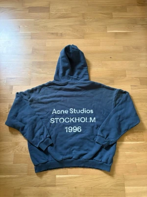 Blå hoodie Acne Studios Stockholm - Mörkblå hoodie från Acne Studios med stor huva och tryck på ryggen där det står 'Acne Studios STOCKHOLM 1996'. Tillverkad i mjuk bomullsblandning och har ribbade muddar vid ärmar och nederkant. Perfekt för en chill och stilren look.