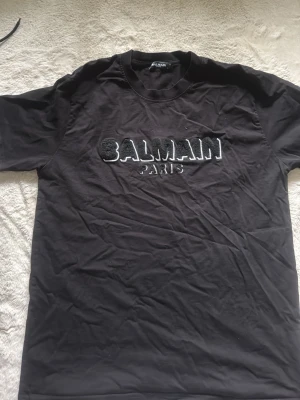 Svart Balmain Paris t-shirt - Säljer en svart t-shirt från Balmain Paris med stor vit och svart broderad logga på bröstet. Klassisk rund halsringning och korta ärmar. Tillverkad i mjuk bomull för skön känsla och stilren look. Storlek Xl men skulle säga att den passar mer som X eller M