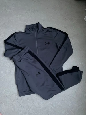  Under Armour - Snyggt grått träningsset från Under Armour med svarta detaljer längs ärmar och ben. Setet består av en långärmad tröja med halv dragkedja och matchande byxor med resår i midjan. Perfekt för gymmet eller löprundan.