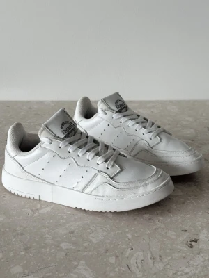 Adidas Originals Supercourt | EU 38 - Snygga vita sneakers från Adidas i äkta läder med klassisk låg siluett. Skorna har perforerade detaljer på sidorna, vit sula och Adidas-logga på plösen och hälen. Perfekta för en clean och stilren look.