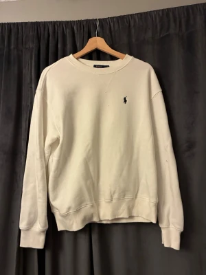 Vit sweatshirt från Polo Ralph Lauren - Klassisk vit sweatshirt från Polo Ralph Lauren med rund halsringning och diskret broderad logga på bröstet. Tröjan har ribbade muddar vid ärmslut och nederkant, perfekt för en clean och avslappnad stil.