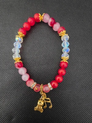 Handgjort pärlarmband med berlocker - Säljer ett handgjort armband med röda, rosa och genomskinliga pärlor. Armbandet har guldfärgade detaljer och två berlocker: en nalle och en musiknot. Perfekt för dig som gillar söta accessoarer med lite bling.