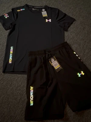 Svart träningsset från Under Armour - Snyggt svart träningsset från Under Armour med t-shirt och shorts. Setet har regnbågsfärgade loggor och textdetaljer på både tröja och byxa. Materialet är lätt och andas, perfekt för gymmet eller löpning. Elastisk midja med dragsko på shortsen.