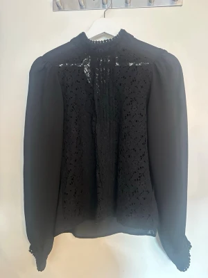 Svart spetsblus med hög krage - Stilren svart blus med transparenta långärmar och detaljer i spets framme. Hög krage med volangkant och knäppning bak i nacken. Blusen har en lätt v-ringning fram och smala spetsdetaljer vid ärmsluten. Perfekt för en trendig och elegant look.