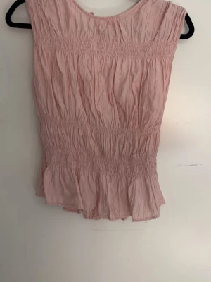 Ljusrosa blus med volang - Söt ljusrosa blus med smockdetaljer och volang nedtill. Blusen är ärmlös och har en rund halsringning. Materialet är tunt och luftigt, perfekt för varma dagar. Passar till både jeans och kjol för en trendig look.