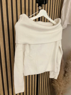 Vit off shoulder stickad tröja - Trendig vit stickad tröja med bred off shoulder-krage och långa ärmar. Tröjan har ribbstickade detaljer vid krage, ärmslut och nederkant. Perfekt för dig som gillar en stilren och modern look.