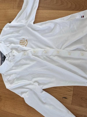 Vit skjorta från Tommy Hilfiger - Klassisk vit skjorta från Tommy Hilfiger med guldigt broderat TH-emblem på bröstet. Skjortan har långa ärmar, knappar framtill och diskreta röda och blå detaljer i kragen. Tillverkad i bomull för en bekväm känsla.