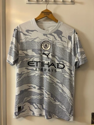 Manchester City bortatröja Puma - Snygg Manchester City bortatröja från Puma i vitt och grått med grafiskt mönster. Tröjan har klubbmärke på bröstet, svart Puma-logga och Etihad Airways som sponsortryck. Tillverkad i lätt och ventilerande funktionsmaterial, perfekt för fotboll eller träning.