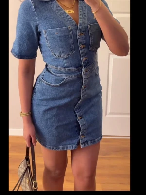 Blå jeansklänning med knappar - Snygg blå jeansklänning med korta ärmar och knappar framtill. Klänningen har två bröstfickor och markerad midja, vilket ger en cool och trendig look. Perfekt för dig som gillar denim och vill ha en casual stil.