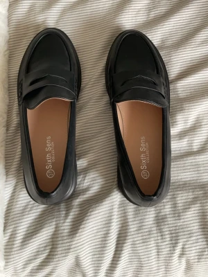 Svarta loafers från Sixth Sens - Säljer ett par klassiska svarta loafers från Sixth Sens. Skorna har chunky sula, stilren pennyloafers-design och är gjorda i slätt konstläder. Insidan är beige och de har en snygg detalj över vristen. Perfekta för dig som gillar en clean och modern look.