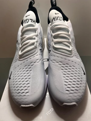 Nike Air Max 270 vita sneakers - Nike Air Max 270 sneakers i vitt med svart detalj på hälen och synlig Air-enhet i sulan. Ovandelen är i mesh för bra ventilation och har tydliga AIR 270-loggor på plösen. Snygg, sportig design med klassisk Nike Swoosh på sidan och tå.