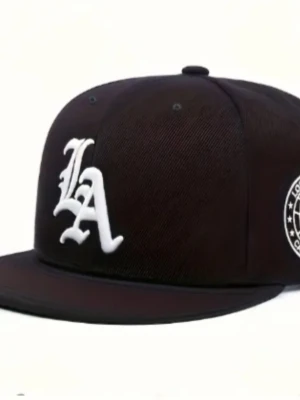 Svart LA snapback keps - Snygg svart snapback keps med stor vit LA-logga broderad framtill och ett runt emblem på sidan. Platt skärm och klassisk form som ger en streetwear vibe. Perfekt för dig som vill ha en stilren och trendig accessoar.