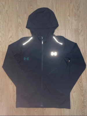 Svart hoodie från Under Armour - Snygg svart hoodie från Under Armour med dragkedja och huva. Tillverkad i funktionsmaterial, perfekt för träning. Reflekterande detaljer på axlar och logga framtill. Modellen heter Threadborne och har en sportig passform.