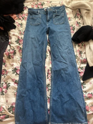 Blå utsvängda jeans Never Denim - Säljer ett par blå högmidjade jeans från Never Denim med utsvängda ben och höga fickor framtill. Jeansen har bälteshällor, dragkedja och knappstängning. Perfekta för dig som gillar en retro och avslappnad stil.